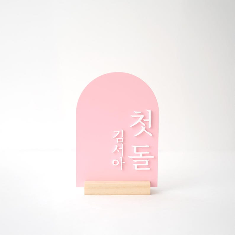 Puede incluir: Un letrero de acr&iacute;lico rosa con una parte superior arqueada y texto coreano blanco. El letrero est&aacute; montado sobre una peque&ntilde;a base de madera rectangular. El fondo es blanco.