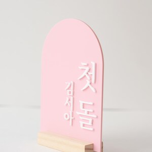 Puede incluir: Un letrero rosa en forma de arco con texto coreano blanco, montado sobre una base de madera natural. La parte superior redondeada del letrero y el contraste entre el rosa y el blanco crean un dise&ntilde;o visualmente atractivo. El letrero probablemente se utiliza para decoraci&oacute;n o anuncios.