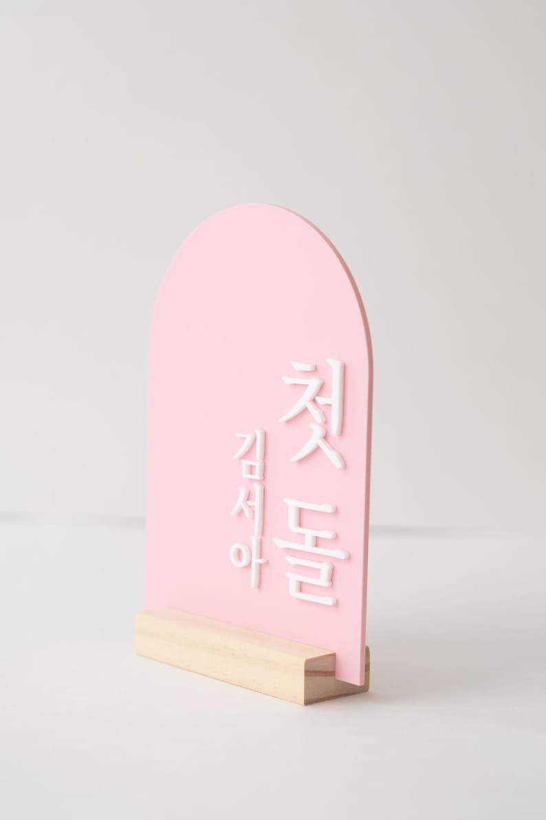 Puede incluir: Un letrero rosa en forma de arco con texto coreano blanco, montado sobre una base de madera natural. La parte superior redondeada del letrero y el contraste entre el rosa y el blanco crean un dise&ntilde;o visualmente atractivo. El letrero probablemente se utiliza para decoraci&oacute;n o anuncios.
