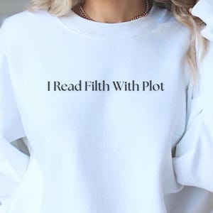 May include: A white sweatshirt with the text "I Read Filth With Plot" in black. The long-sleeved crew neck sweatshirt is made of a soft material. The sweatshirt is a casual style.