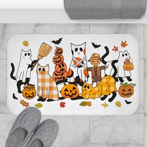 Op de afbeelding: Witte rechthoekige Halloween-badmat met cartoonkatten verkleed als geesten, vogelverschrikkers en pompoenen. Het ontwerp bevat pompoenen, vleermuizen en herfstbladeren. Een paar grijze slippers staan op de voorgrond.