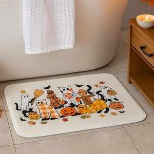 Puede incluir: Una alfombra de baño rectangular con temática de Halloween con gatos de dibujos animados disfrazados, calabazas, murciélagos y hojas de otoño. La alfombra es de color crema y está colocada sobre un suelo embaldosado cerca de una bañera blanca y un mueble de madera.