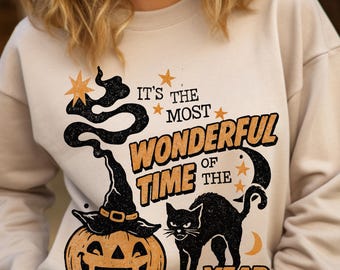 May include: Beige sweatshirt with a Halloween-themed graphic. The design includes a jack-o'-lantern, a black cat, a witch's hat, and the text "It's the Most Wonderful Time of the Year" in orange and black.