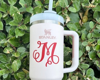 May include: A white Stanley travel mug with a red monogram "M" and a straw. The mug has a handle and a stainless steel lid.