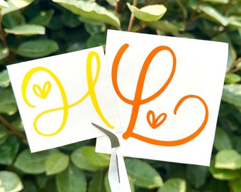 May include: Two white square stickers with the letters "H" and "L" in yellow and orange, respectively. Each letter has a small heart detail. The stickers are held by tweezers against a backdrop of green foliage.