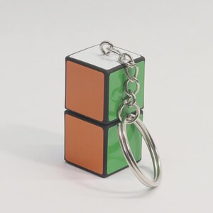 May include: A two-layered Rubik's Cube keychain. The top layer has white, orange, and green sides, while the bottom layer has orange and green sides. A silver chain and keyring are attached to the top.