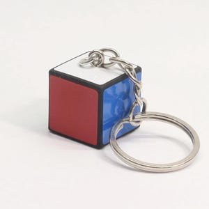 Op de afbeelding: Een Rubik's Cube sleutelhanger met een rood, wit en blauw ontwerp. De kleine kubus is bevestigd aan een zilverkleurige ketting en sleutelring. De sleutelhanger is een miniatuurversie van het klassieke puzzelspel.
