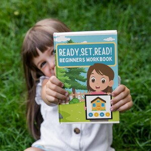 May include: A beginner's workbook titled "READY, SET, READ!" is held up in front of a green grassy background. The cover features a cartoon illustration of a girl and a house, with a blue and green border.