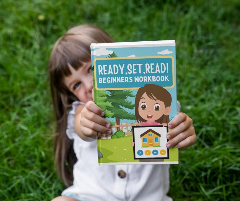 May include: A beginner's workbook titled "READY, SET, READ!" is held up in front of a green grassy background. The cover features a cartoon illustration of a girl and a house, with a blue and green border.