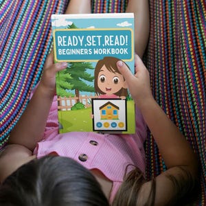 May include: A child holding a "READY, SET, READ! BEGINNERS WORKBOOK" book. The book cover features a cartoon girl and a house illustration. The child is wearing a pink shirt and is lying in a colorful hammock.