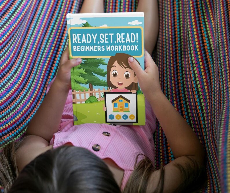 May include: A child holding a "READY, SET, READ! BEGINNERS WORKBOOK" book. The book cover features a cartoon girl and a house illustration. The child is wearing a pink shirt and is lying in a colorful hammock.
