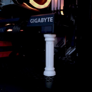 Może przedstawiać: Miniaturowa biała kolumna z żłobionym trzonem i kwadratową podstawą. Kolumna stoi na czarnej powierzchni z komputerowym komponentem w tle.