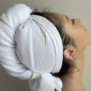 May include: A white microfiber hair towel with a white elastic band. The towel is wrapped around a person's head, with the elastic band securing it in place.