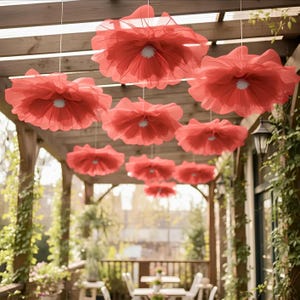 May include: Red paper flower decorations hanging from a wooden pergola. The flowers have a layered petal design with a white center. The outdoor setting includes a patio with tables and chairs.
