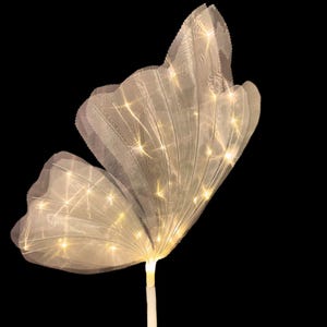 Op de afbeelding: Een wit, doorschijnend, vlindervormig licht met warm witte LED-verlichting. Het licht bevindt zich op een witte paal.