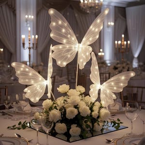 Puede incluir: Centro de mesa con tres decoraciones de mariposas iluminadas. Cada mariposa tiene alas blancas iluminadas con pequeñas luces. Están dispuestas sobre un lecho de rosas blancas y vegetación, creando una elegante exhibición.
