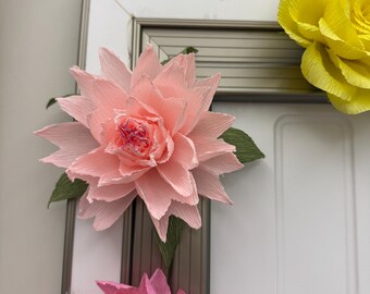 May include: Close-up of handmade paper flowers in shades of pink and yellow. The pink flower is in the foreground, with green leaves and a detailed center. A yellow flower is visible in the background. The flowers are attached to a silver frame.