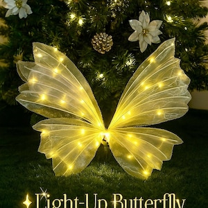 Könnte beinhalten: Leuchtende Schmetterlingsdekoration für Weihnachten. Der Schmetterling hat gelbe Flügel mit kleinen eingebetteten Lichtern, vor dem Hintergrund eines geschmückten Weihnachtsbaums. Der Text "Light-Up Butterfly - Decor - Christmas Edition" steht unten.