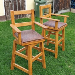 May include: Two wooden bar stools with brown leather seats and a simple design. The stools are made of natural wood and have a rustic look.