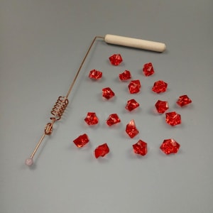 May include: A copper-colored tool with a wooden handle and a coiled spring, next to scattered red, faceted, gem-like pieces. A small, pink, round bead is at the end of the tool.
