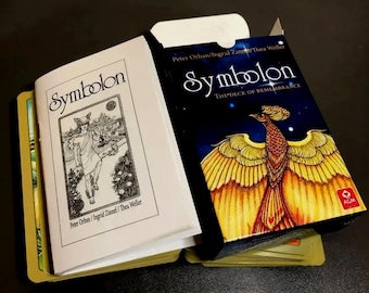 May include: A closed book with the title "Symbolon: The Deck of Remembrance" on the cover. The cover features a golden phoenix with a blue background and stars. The book is open to reveal a black and white illustration of a woman in a forest.