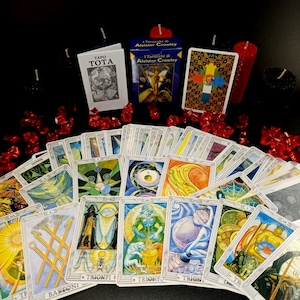 Può includere: Uno spargimento di carte dei tarocchi con illustrazioni colorate su sfondi bianchi. Le carte sono disposte a ventaglio con il testo "I Tarocchi di Aleister Crowley" visibile sulla carta superiore. Le carte sono circondate da candele rosse e nere e da dadi rossi.