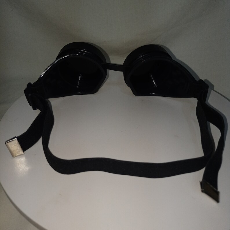 May include: Black goggles featuring two circular, dark-tinted lenses connected by a bridge. The frame is constructed from a solid black material, and the eyewear includes an adjustable black elastic strap with silver-toned metal end fasteners.