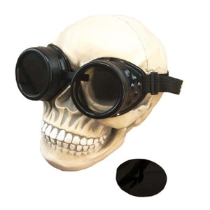 May include: A realistic off-white human skull model wearing black circular goggles with dark lenses and an adjustable elastic strap. A separate, detached circular black lens is positioned below the skull on the surface.