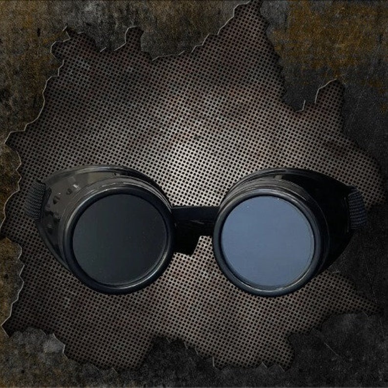 May include: Black goggles featuring two circular, dark-tinted lenses encased in a solid frame. The eyewear includes a black adjustable elastic strap attached to the sides, designed to fit securely around the head for eye protection and coverage.