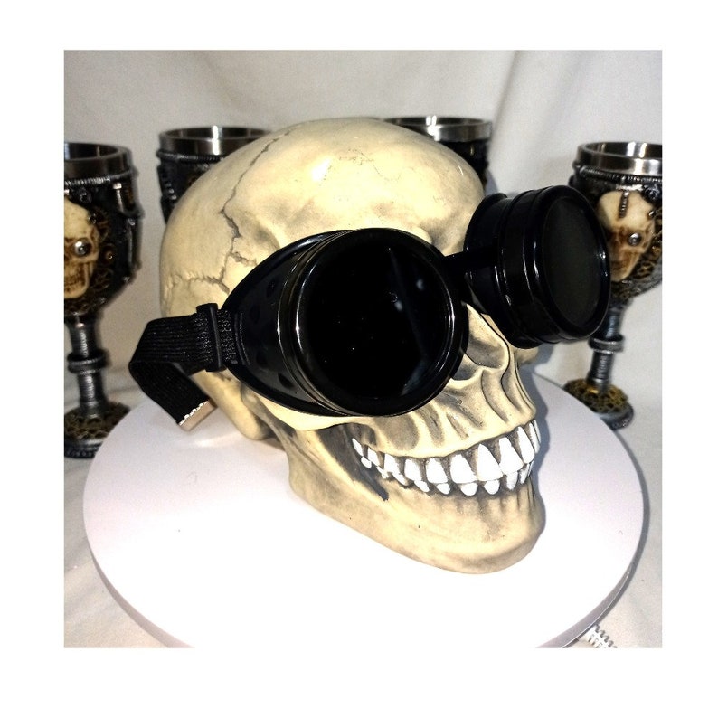 May include: A beige human skull replica wearing black circular goggles with dark lenses and an adjustable black elastic strap. The skull features realistic bone-colored textures, defined sutures, and white teeth, positioned with goggles over the eye sockets.