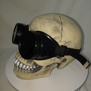 May include: A beige human skull model featuring realistic bone texture and sutures, wearing a pair of black plastic goggles with dark lenses and an adjustable black elastic strap secured around the cranium, presented in a side profile view.