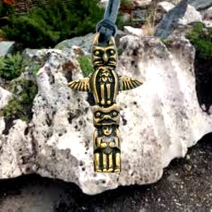 Peut inclure: Un pendentif en métal doré représentant un totem avec un hibou en haut, une figure humaine au milieu et un oiseau en bas. Le pendentif est suspendu à un cordon en cuir noir.