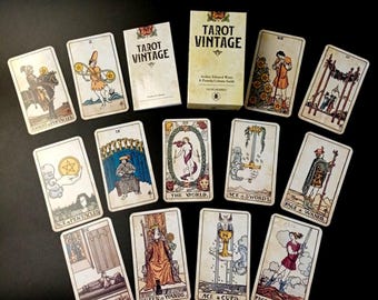 May include: Tarot Vintage deck of cards spread out on a black surface. The cards feature illustrations of various tarot symbols, including the World, Ace of Swords, Page of Wands, Queen of Wands, Ace of Cups, and Page of Swords. The cards are in a vintage style with a cream background and brown and black illustrations.