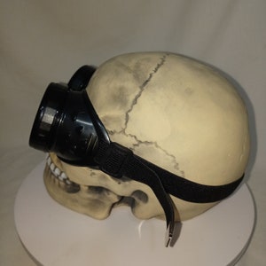 May include: A beige human skull model wearing black plastic goggles with a black elastic strap. The goggles feature circular lenses and side vents, positioned over the eye sockets of the skull, which shows detailed bone texture and sutures.