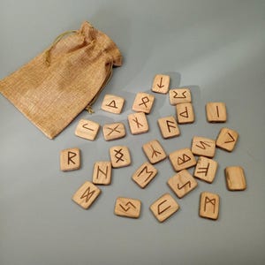 May include: A set of wooden runes in a burlap bag. The runes are engraved with Norse symbols and are arranged on a gray surface.