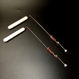 Op de afbeelding: Twee koperkleurige metalen gereedschappen met witte keramische handvatten en spiraalveren. Elk gereedschap heeft een kleine, heldere, ronde kraal aan het einde van een dunne draad. De gereedschappen zijn schuin en liggen op een zwart oppervlak.