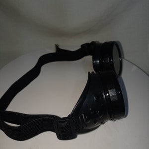 May include: Black goggles featuring circular lenses and a side-mounted ventilation design. The eyewear includes a flexible, adjustable black elastic strap with a plastic buckle, designed to fit securely around the head for eye protection.