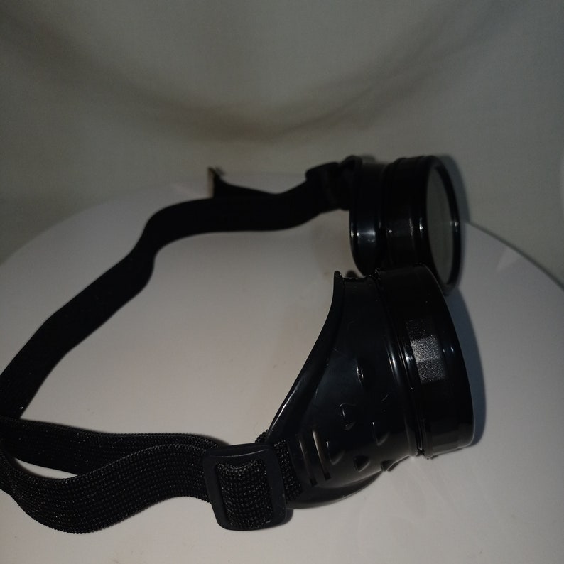 May include: Black goggles featuring circular lenses and a side-mounted ventilation design. The eyewear includes a flexible, adjustable black elastic strap with a plastic buckle, designed to fit securely around the head for eye protection.