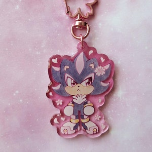 May include: A pink acrylic keychain with a cartoon character in blue and white, red eyes, and a serious expression. The keychain has a rose gold flower-shaped clasp and is decorated with small hearts and flowers.