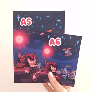 May include: Two art prints featuring a character from the Sonic the Hedgehog series. The prints are labeled "A5" and "A6" and depict the character in a city scene under a starry night sky. The color palette includes shades of red, black, and blue.