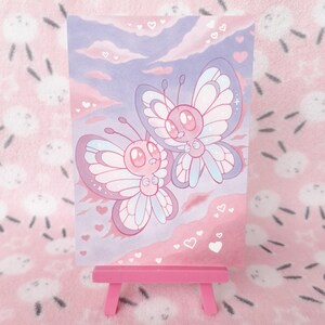 May include: A pink and purple illustration of two cartoon butterflies with heart accents. The artwork is displayed on a small pink easel. The background features a pink and purple sky with clouds and hearts.