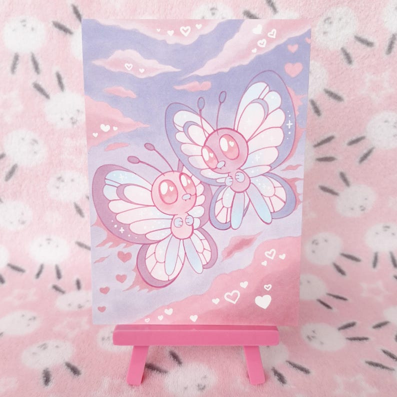May include: A pink and purple illustration of two cartoon butterflies with heart accents. The artwork is displayed on a small pink easel. The background features a pink and purple sky with clouds and hearts.