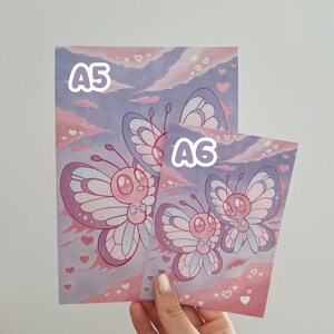 May include: Two art prints with cartoon butterflies in pink, purple, and white. The prints are labelled "A5" and "A6" in white text. The background has a sky design with hearts.