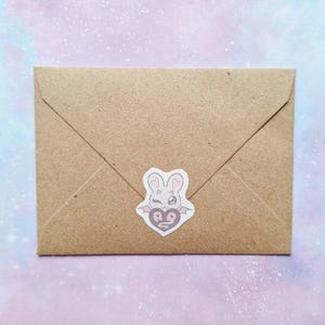 May include: A brown paper envelope with a sticker featuring a bunny head with heart-shaped eyes and bat wings. The sticker is white with pink and gray accents. The envelope is sealed.