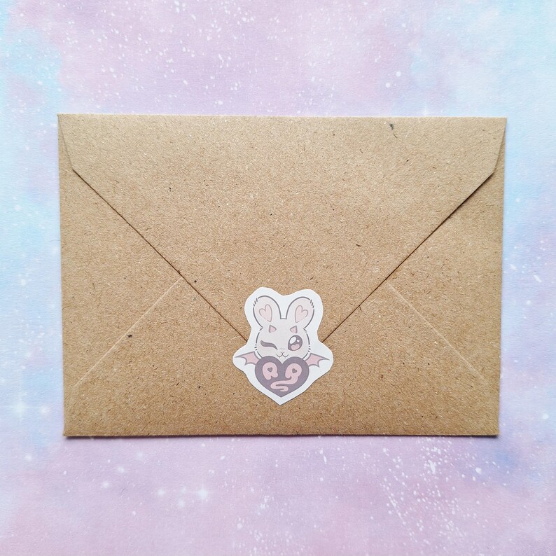 May include: A brown paper envelope with a sticker featuring a bunny head with heart-shaped eyes and bat wings. The sticker is white with pink and gray accents. The envelope is sealed.