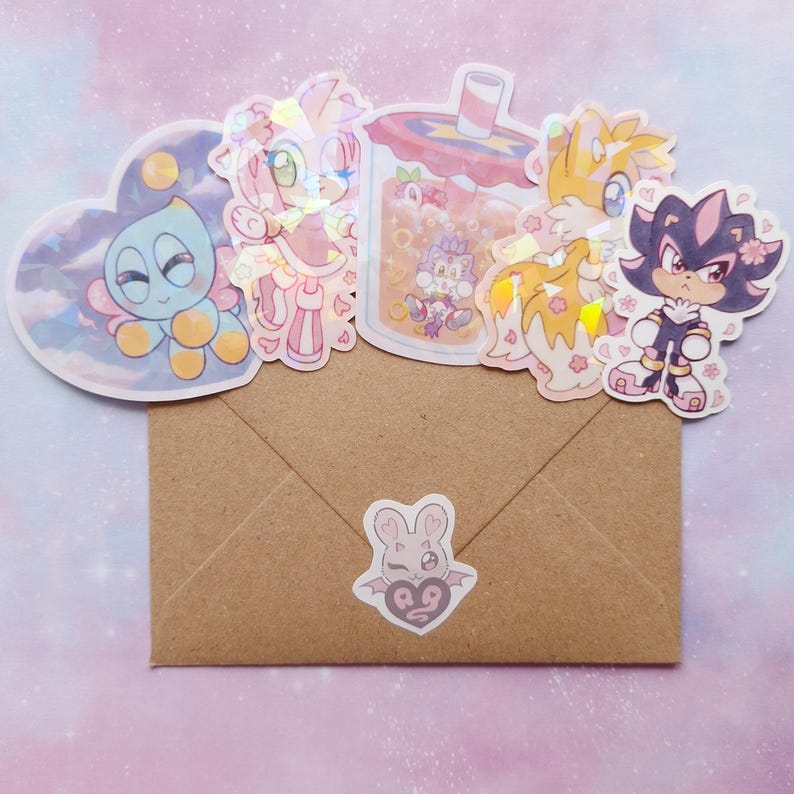 May include: A brown envelope with several holographic stickers of cartoon characters. The stickers include a blue creature, a pink character, a drink, a yellow fox, and a black and purple character. A small sticker of a bunny is on the envelope.