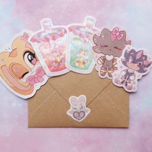 May include: A collection of colorful stickers featuring cartoon characters and bubble tea designs. The stickers are arranged on a brown envelope, with a small sticker of a bat with a heart on the front. The stickers are in various shapes and sizes.