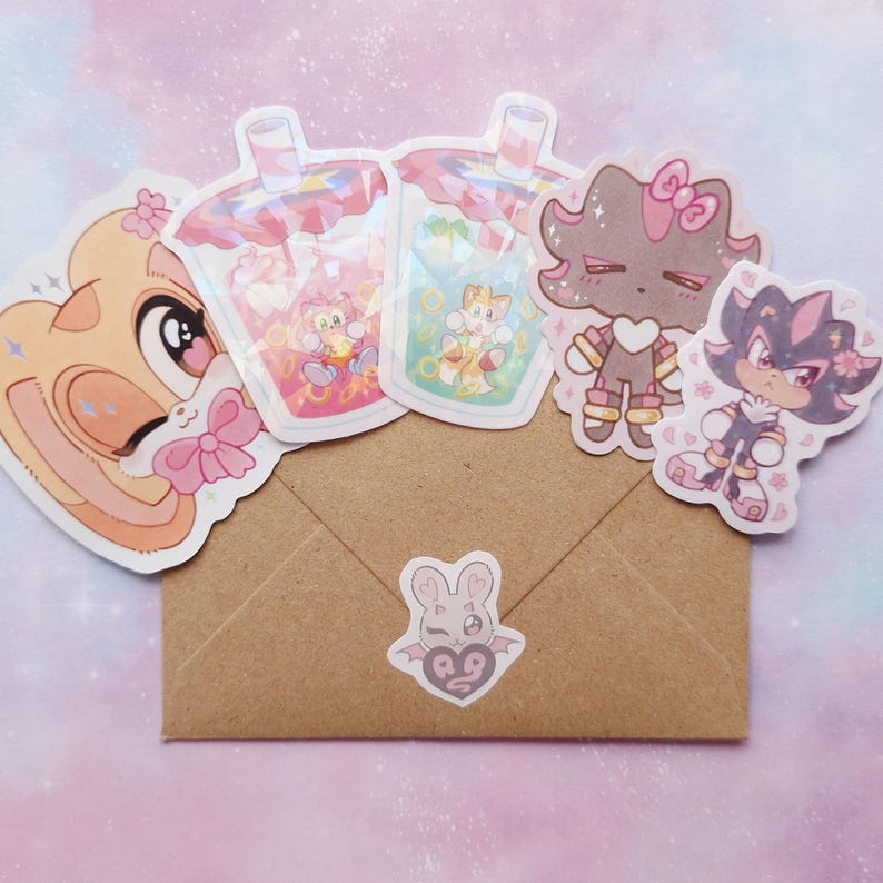 May include: A collection of colorful stickers featuring cartoon characters and bubble tea designs. The stickers are arranged on a brown envelope, with a small sticker of a bat with a heart on the front. The stickers are in various shapes and sizes.