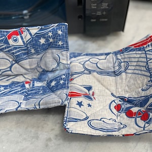 May include: Two blue and white fabric pot holders with a red, white, and blue airplane design. The pot holders are folded and have a quilted texture.