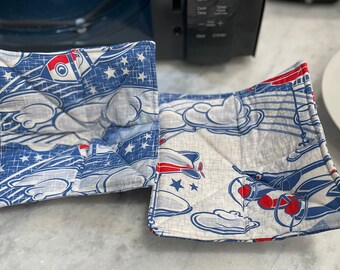 Peut inclure: Deux maniques en tissu bleu et blanc avec un motif d'avion rouge, blanc et bleu. Les maniques sont pliées et ont une texture matelassée.